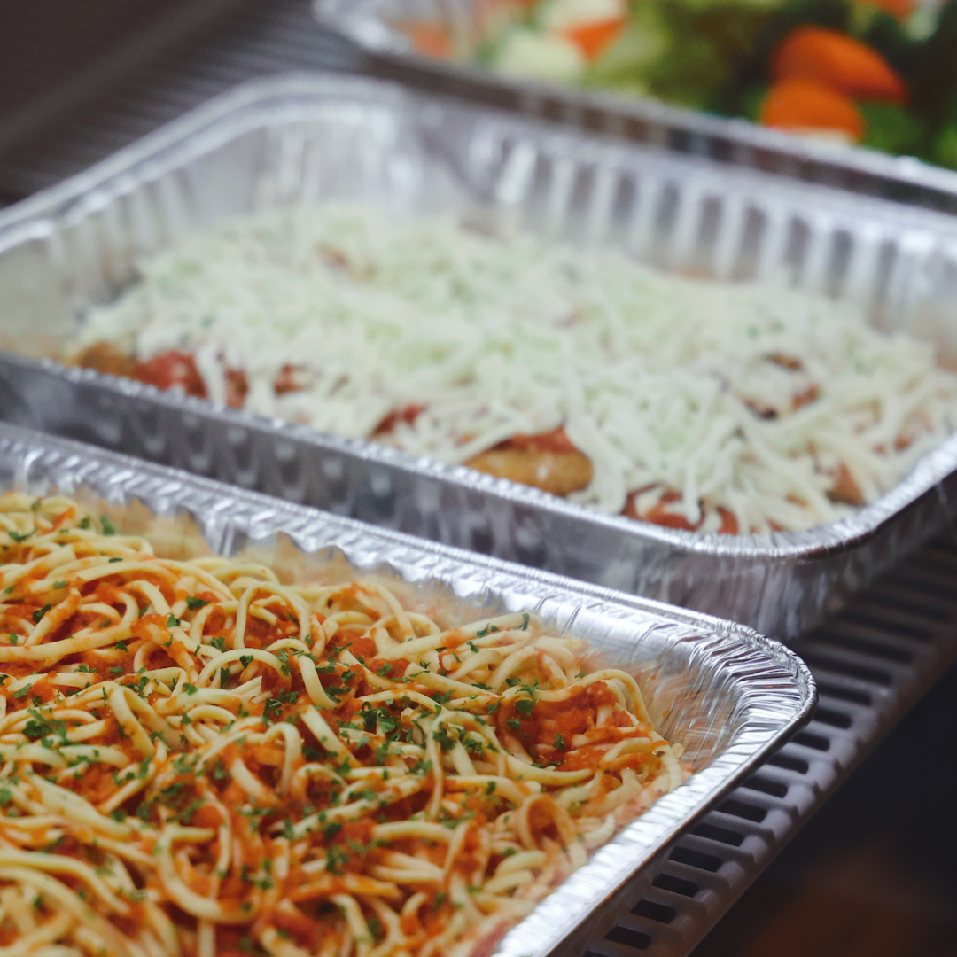 Three aluminum trays of food on a table, featuring spaghetti with sauce and herbs, chicken parmesan with cheese, and mixed vegetables.
