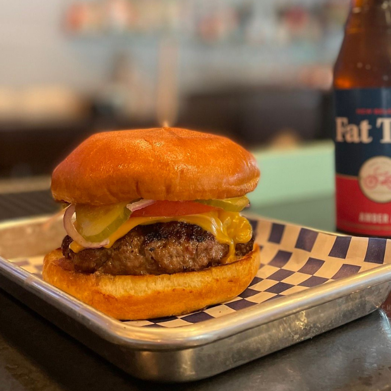 A juicy hamburger with tomatoes, lettuce, and pickles on a brioche bun, served on a tray beside a bottle of fat tire amber ale.