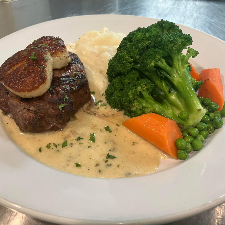 A plate of food with steak topped with a scallop, mashed potatoes, broccoli, carrots, and peas, garnished with herbs in a creamy sauce.
